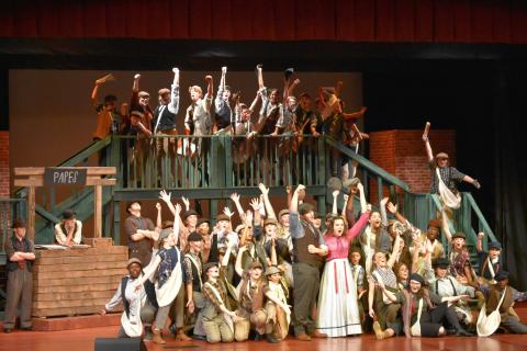 Large cast of Newsies performing on stage with multi-level wooden set, stairs, and railings.