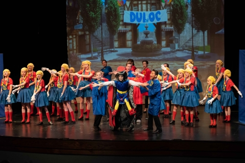 A group of performers in red tops and blue skirts with a performer in the middle wearing a blue velvet tunic in front of a digital projection of a town with a banner that reads "Duloc" in blue letters.