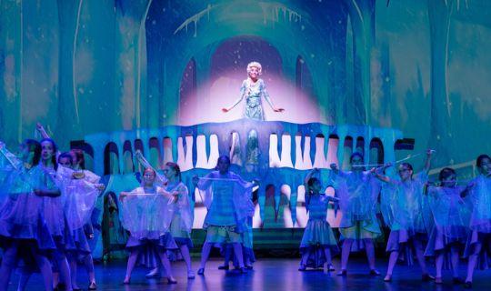 A projection of an ice castle with a girl in a blue dress on an icicle decorated balcony with a group of kids in front.