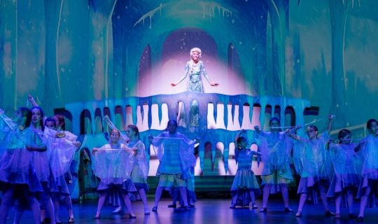 A projection of an ice castle with a girl in a blue dress on an icicle decorated balcony with a group of kids in front.
