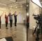Four dancers stand in a row with their arms in their air and feet together in front of a filming camera.