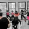 Five dancers wearing pink tops and shorts in a row demonstrating a dance move while someone is filming in front of them.